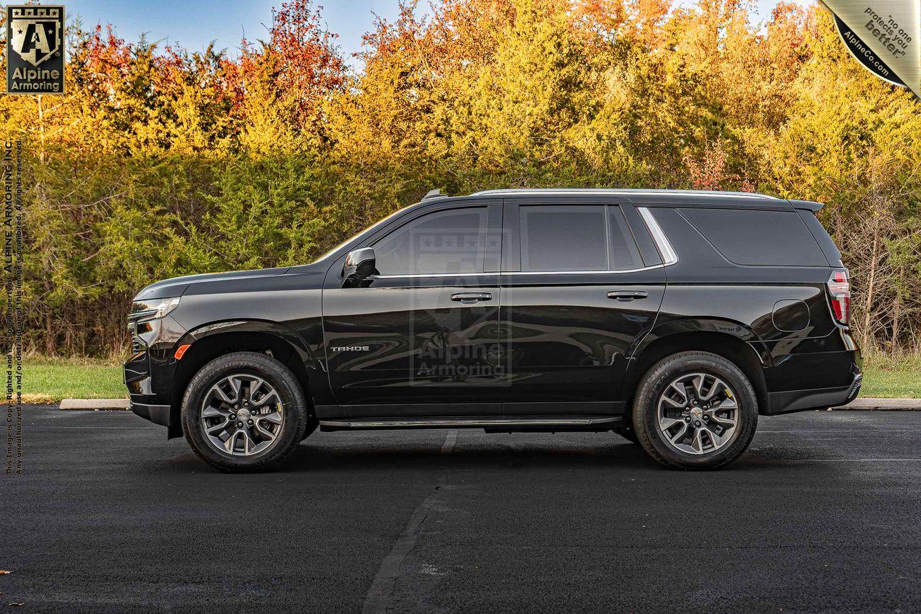 A black Chevrolet Tahoe SUV is parked on a paved surface in front of a line of autumn trees. It has tinted windows, chrome wheels, and is branded with the Alpine Armoring logo.