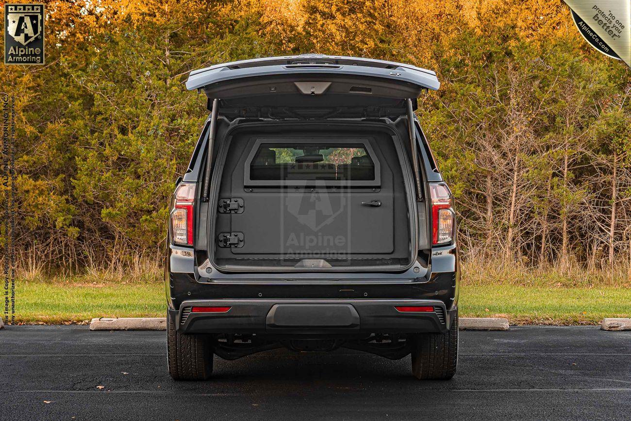 A rear view of an armored black Chevrolet Tahoe SUV with its back door open, showcasing an enclosed storage area with a compartmentalized secure container inside. The background includes trees and a grassy area.