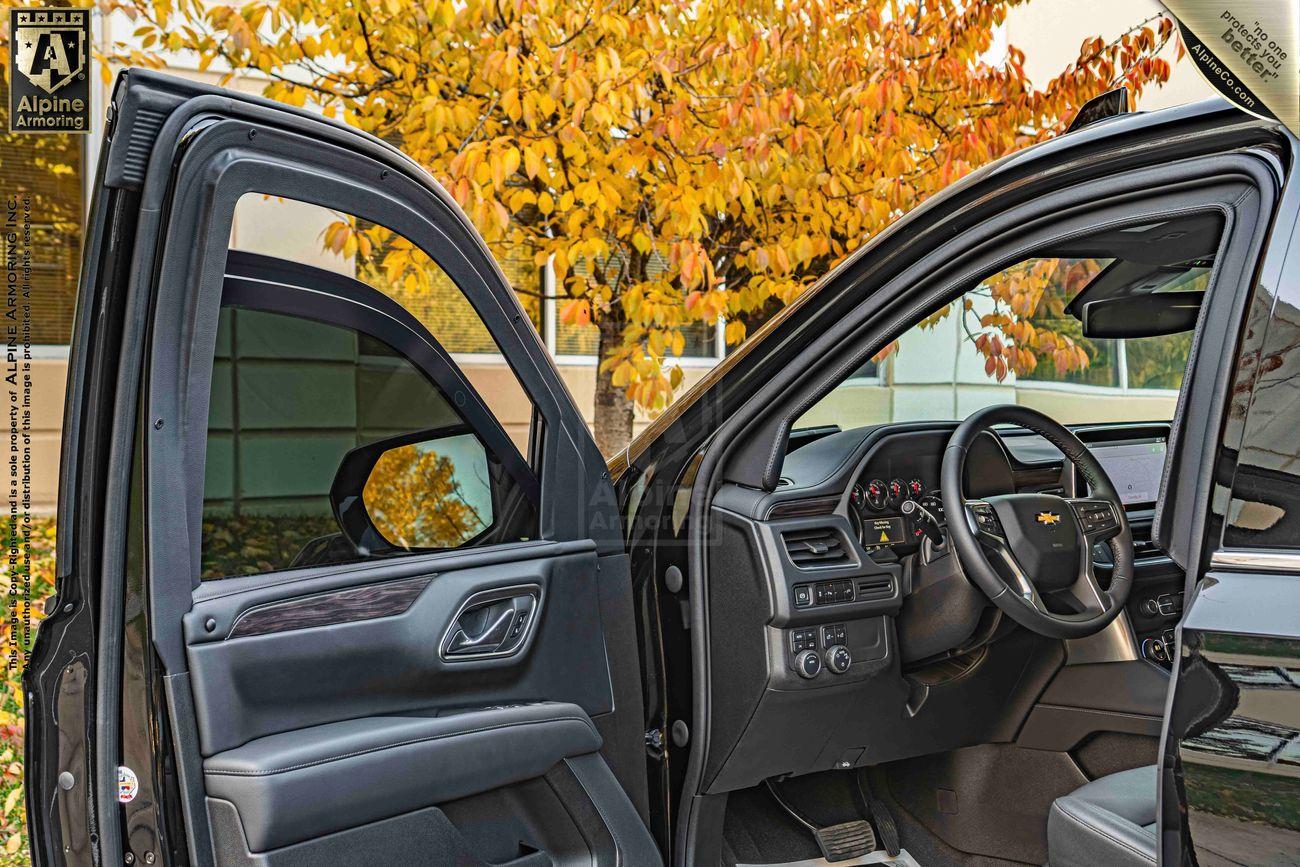 Interior view of an open car door showing the driver's seat, steering wheel, and dashboard of a black Chevrolet Tahoe SUV with autumn foliage visible outside.