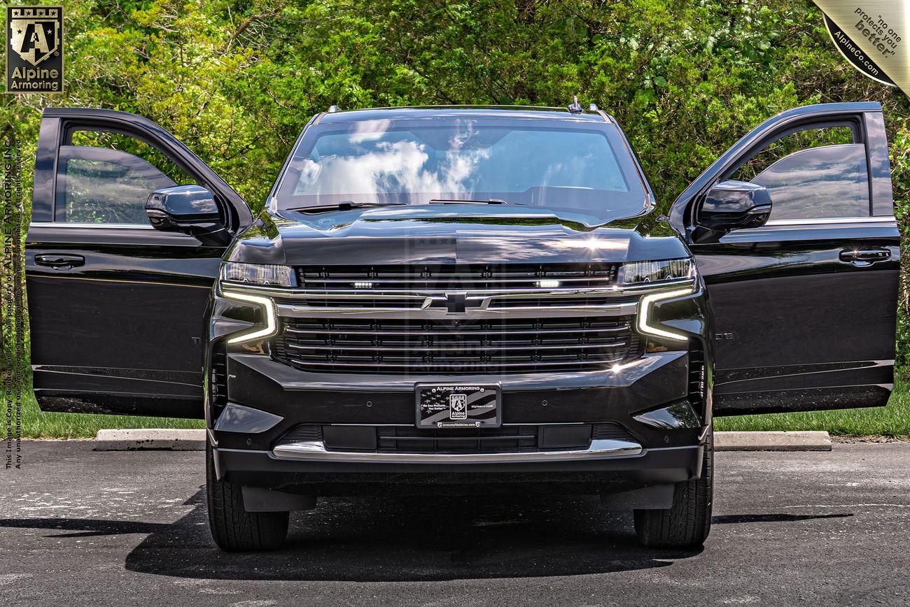 A black bulletproof Chevrolet Tahoe SUV is parked with both front doors open, showing a frontal view against a backdrop of green trees and a partly cloudy sky. The vehicle features illuminated LED headlights.