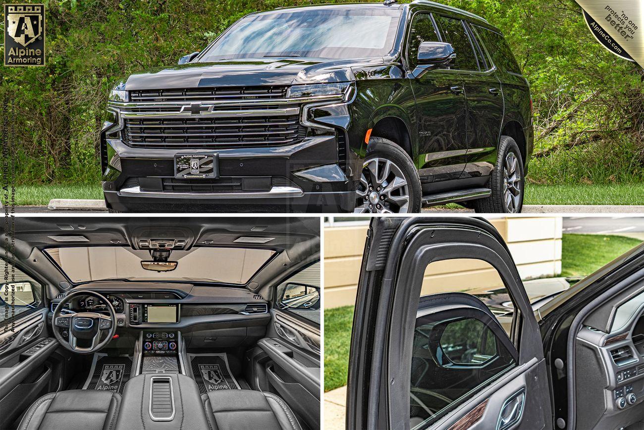 A black, armored Chevrolet Tahoe SUV is shown from the front, the interior features including the dashboard and seats, and the rear door slightly open. The vehicle is displayed against a backdrop of greenery.