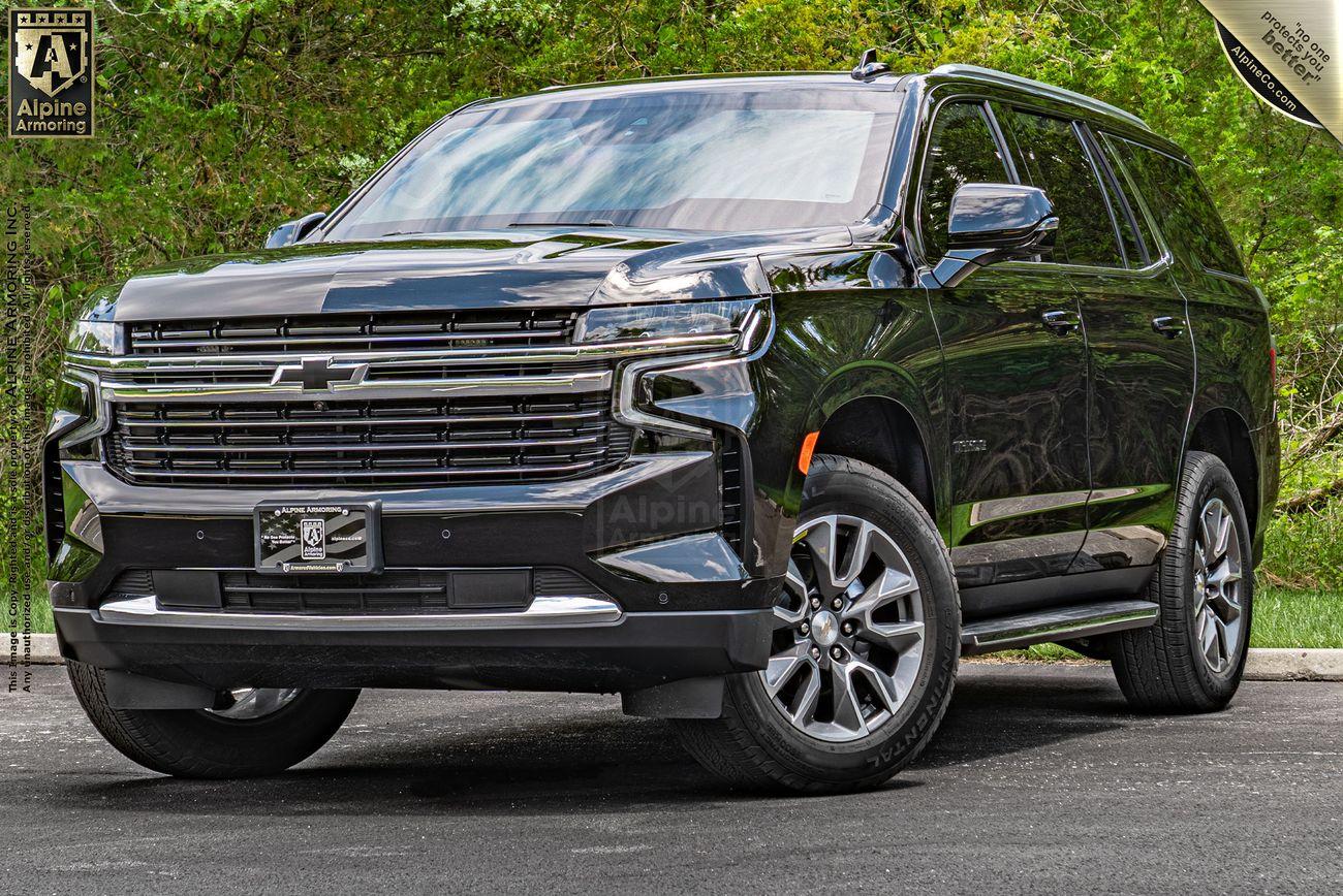 A black, bulletproof Chevrolet Tahoe SUV is parked on a paved surface with greenery in the background. The vehicle has a grille with the Chevrolet emblem and chrome accents on the wheels.