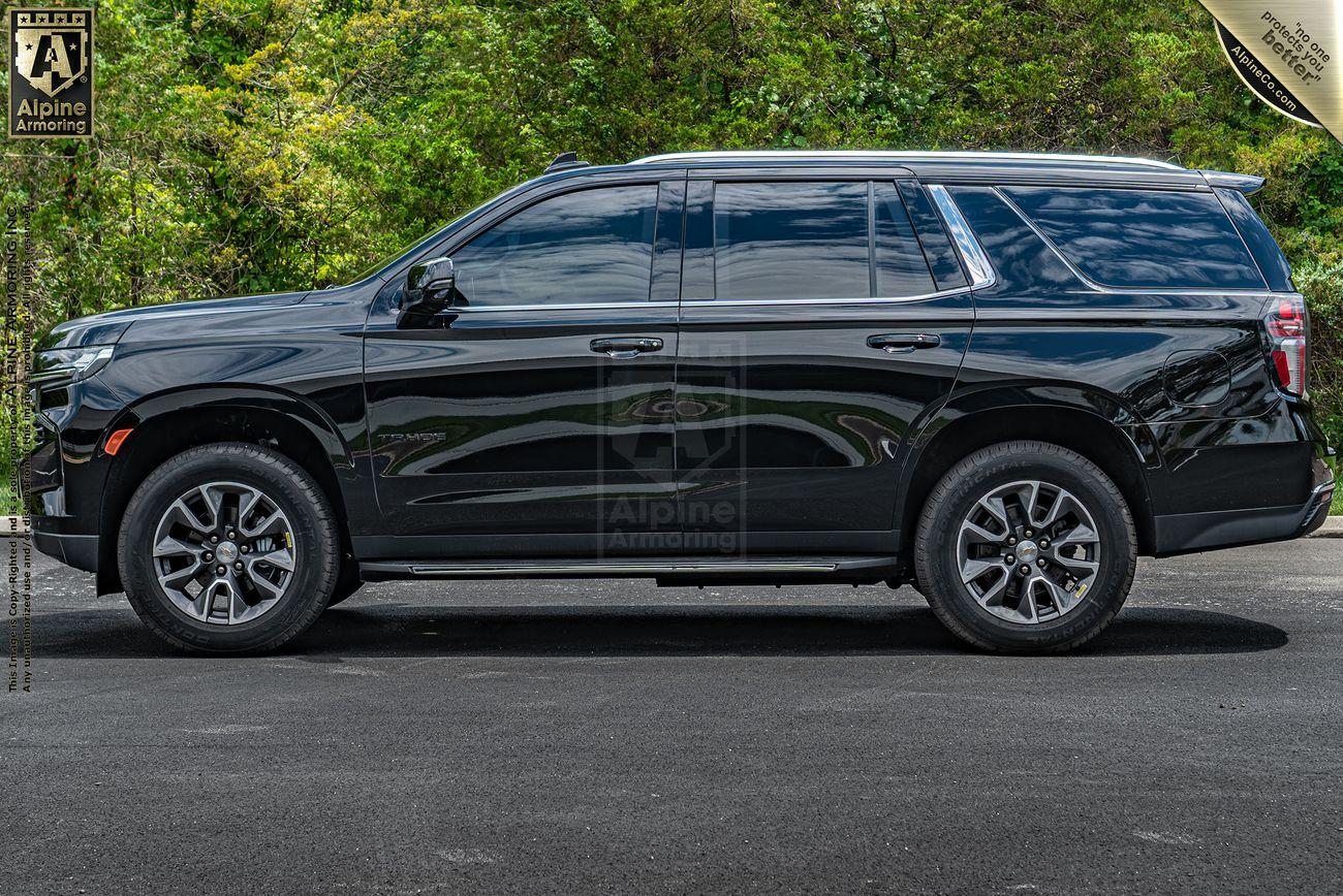 A side view of a black Chevrolet Tahoe SUV parked on a paved surface with greenery in the background. The SUV has tinted windows and alloy wheels.