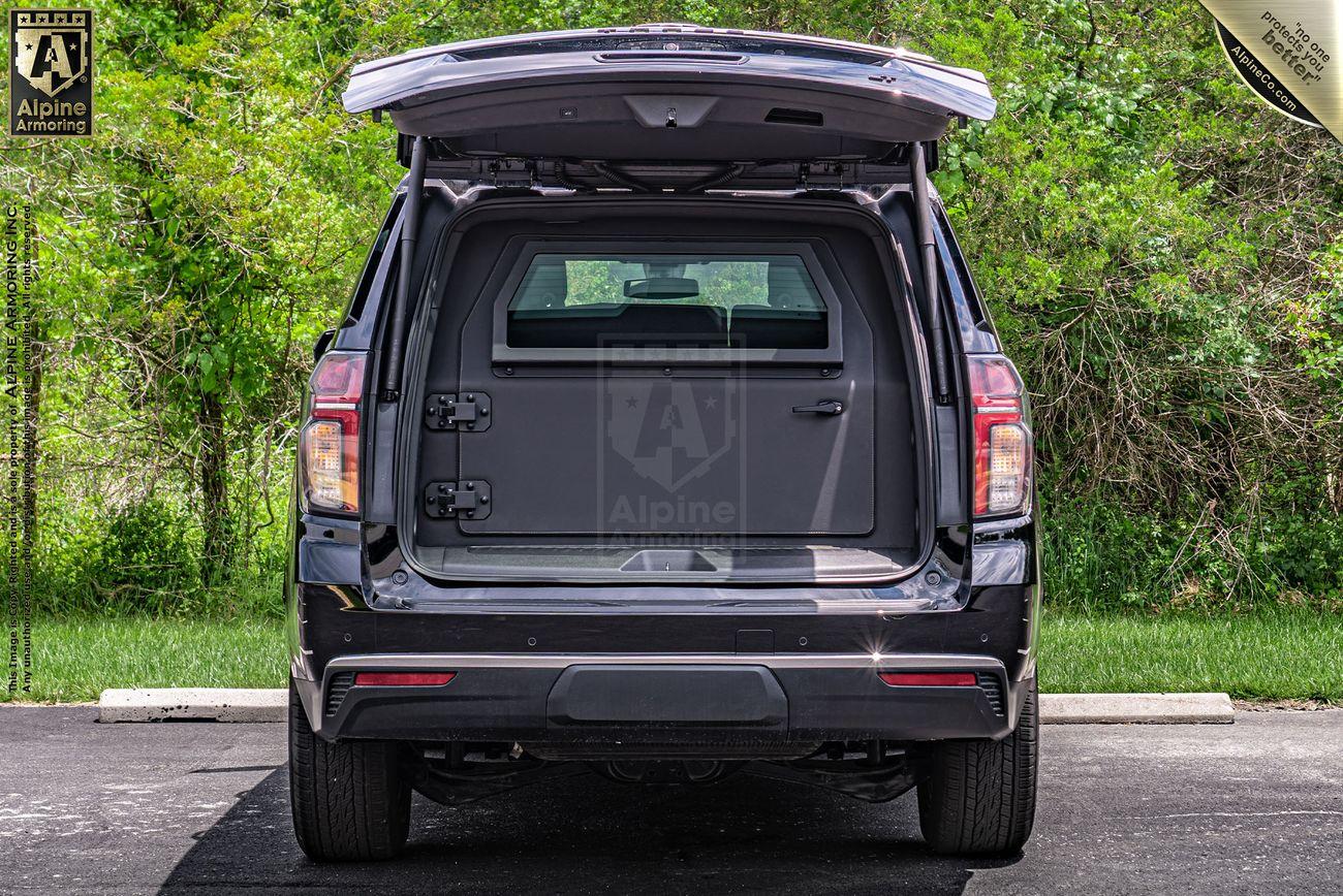 A rear view of an open armored Chevrolet Tahoe SUV trunk with an exterior storage compartment visible. The trunk door is lifted, revealing an interior storage area against a backdrop of greenery.