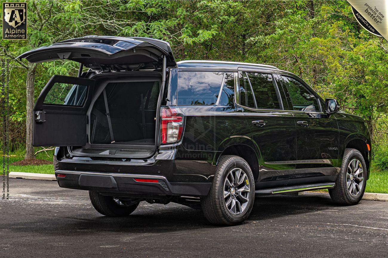 A black Chevrolet Tahoe SUV with its rear hatch open, revealing the cargo space, is parked on a road with trees in the background.