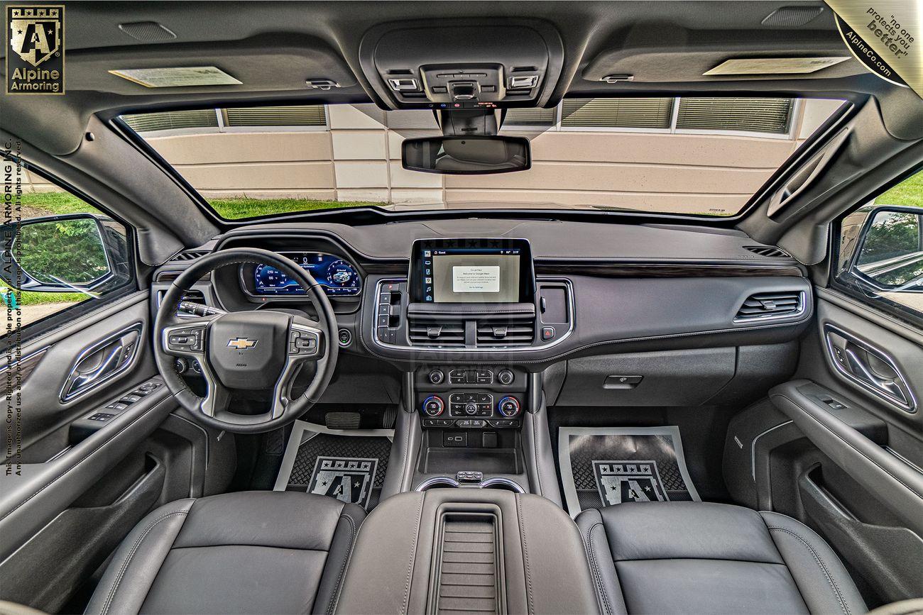 Interior view of an armoredf Chevrolet Tahoe SUV showing the steering wheel, dashboard with digital displays, center console, and leather seats. The dashboard features the Alpine Armoring branding.