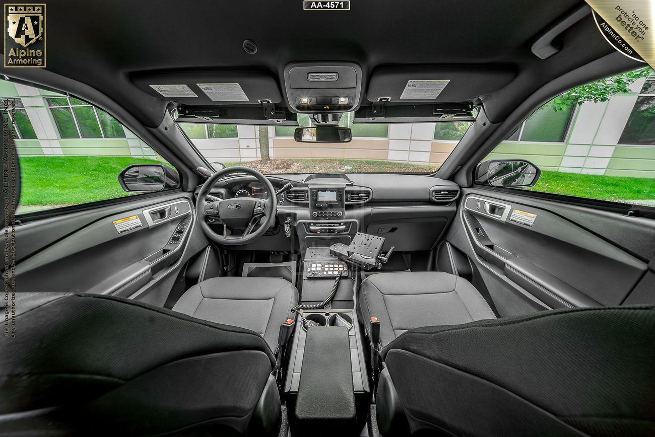 Interior view of an armored police Ford Explorer SUV showing the front seats, dashboard, and various electronic equipment. Greenery and a building are visible through the windows.