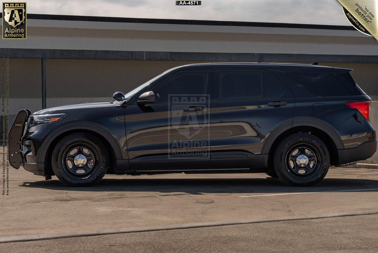 A side view of a black bulletproof Ford Explorer PPV SUV parked on a paved surface in front of a building. The vehicle has a bull bar on the front. The background is a gray structure with a corrugated roof.