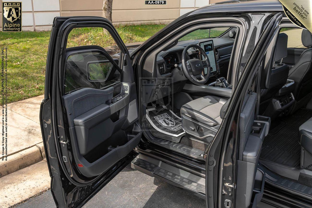 Interior of an armored Ford Lightning with the driver's door open, showcasing the dashboard, steering wheel, and driver seat. There is a logo on the floor mat and a label on the upper part of the door.