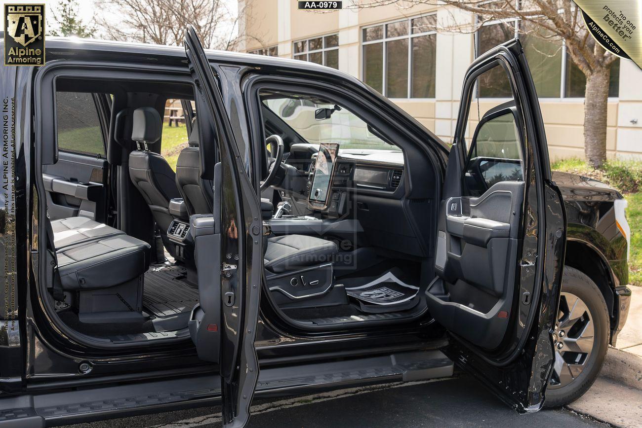 A black armored Ford Lightning with all doors open showcasing a spacious interior with black seats and modern dashboard. The setting is an outdoor parking area near a building.