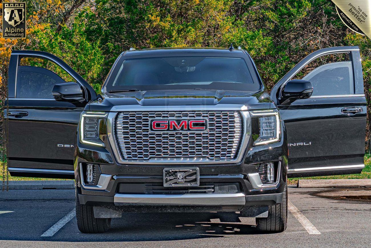 A black GMC Yukon Denali SUV is parked in a parking lot with both front doors open. The SUV features a prominent front grille and is situated in front of green shrubbery on a sunny day.