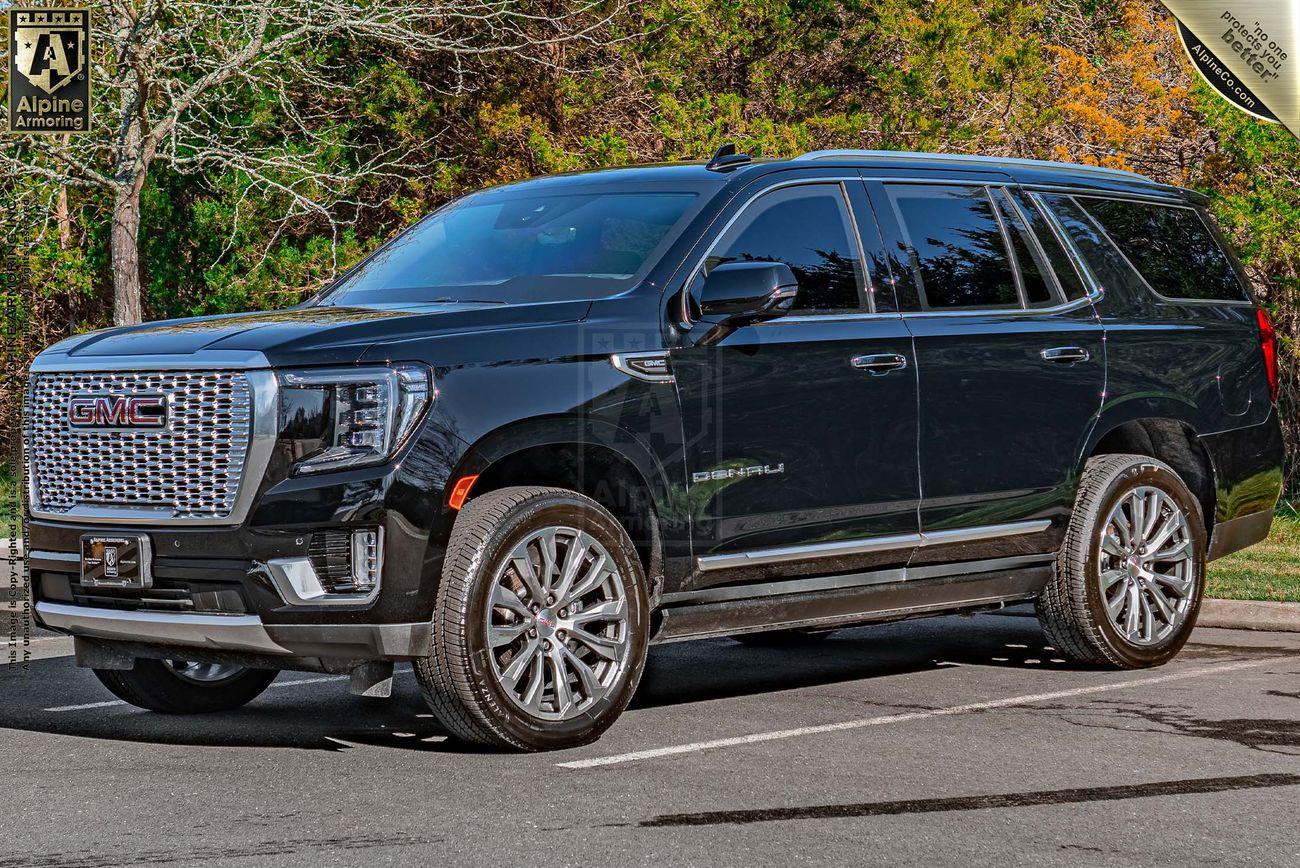 A black bulletproof GMC Yukon Denali SUV is parked in an outdoor setting, surrounded by greenery and trees. The car is facing slightly to the right, showing its side and front grille.