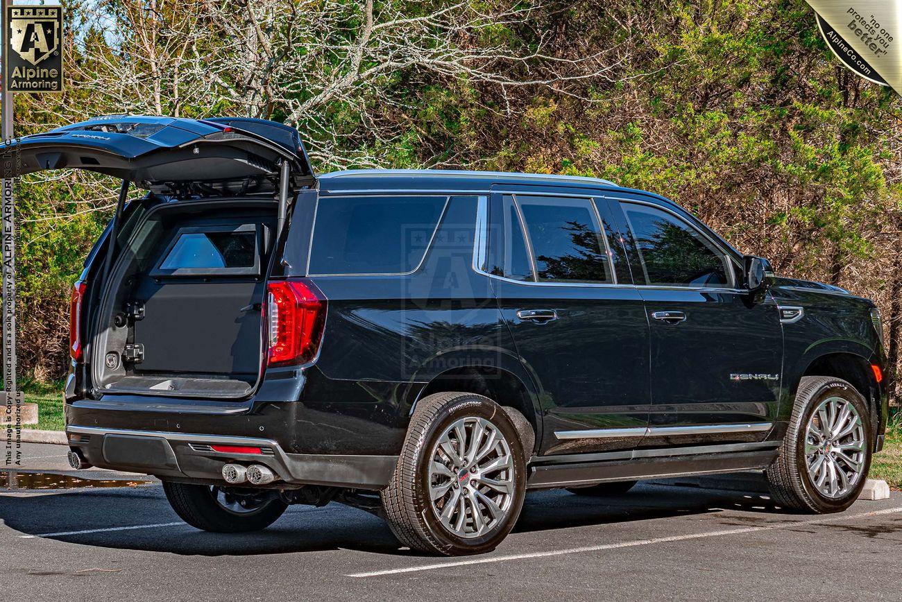 Black armored GMC Yukon Denali SUV with its rear hatch open, parked in an outdoor lot. Background includes trees and some signage on a pole.