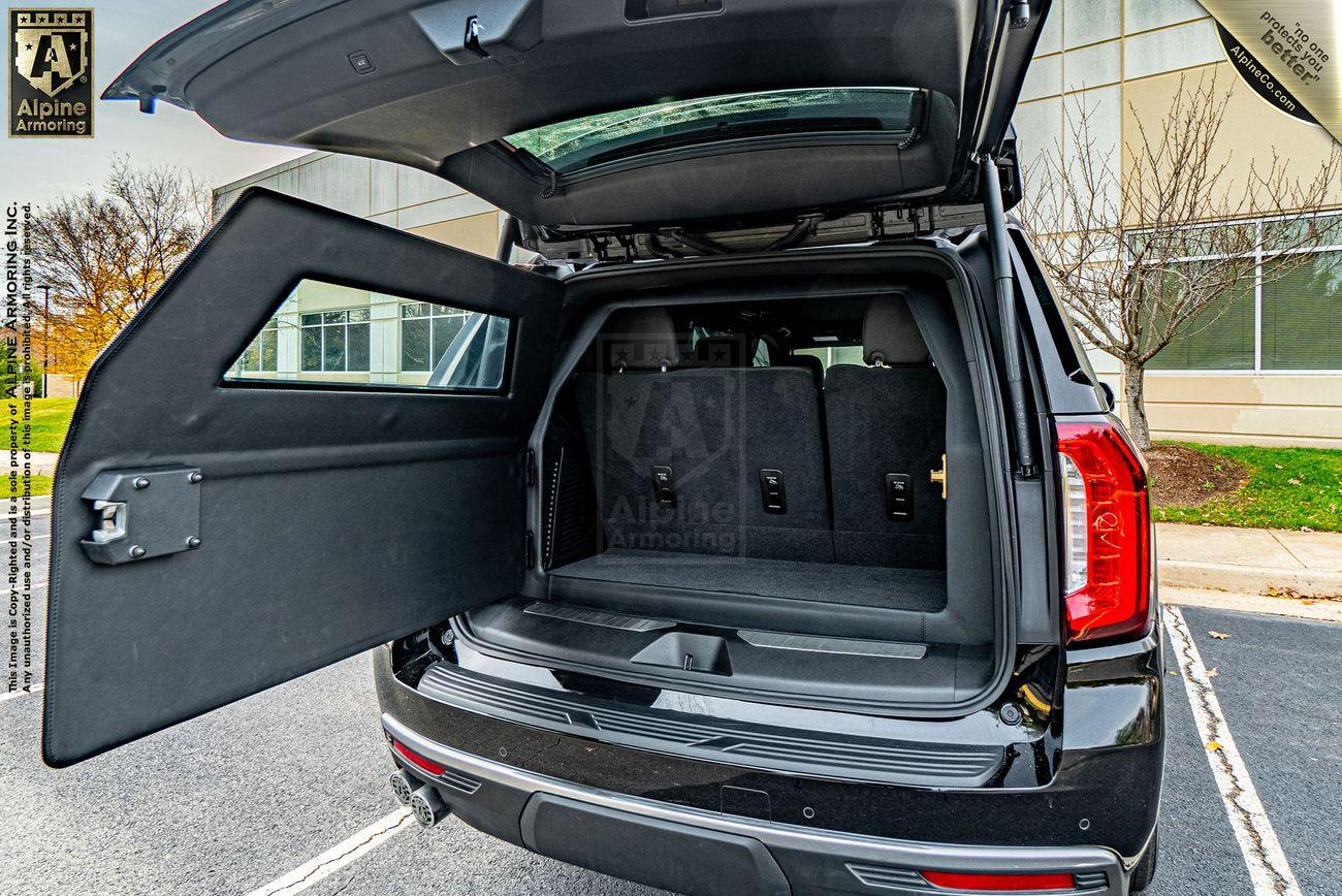 Open rear hatch of a black Lexus LX600 Luxury car showing an armored security compartment with a window cutout. The vehicle is parked outside a modern building.