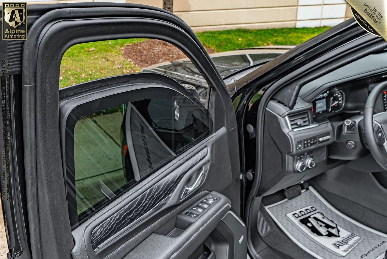 Interior view of an open armored GMC Yukon Denali door highlighting the bulletproof window, window controls, dashboard, and branded floor mat with "Alpine" visible.