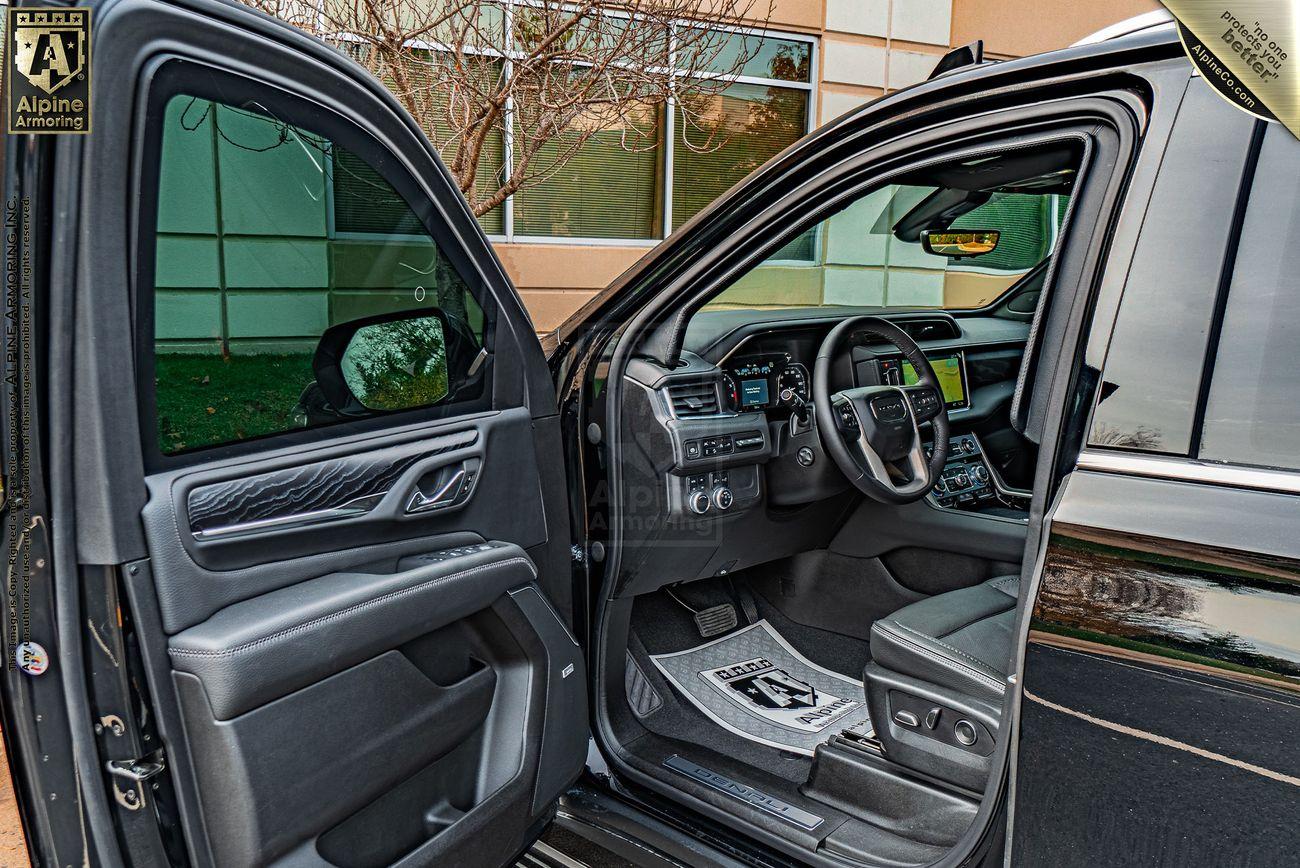 Interior view of an armored GMC Yukon Denali showcasing the driver's seat, dashboard, and steering wheel. The door is open, revealing the controls and materials used in the vehicle's design.