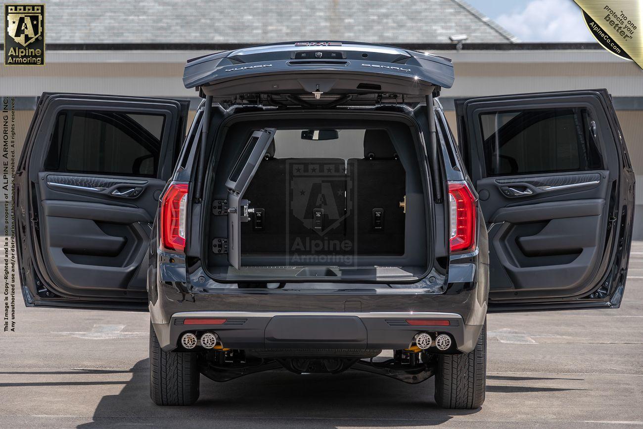 A black armored GMC Yukon Denali XL SUV with all doors and tailgate open, revealing a spacious and empty interior. The vehicle is parked on a concrete surface in front of a building with a sloped roof.