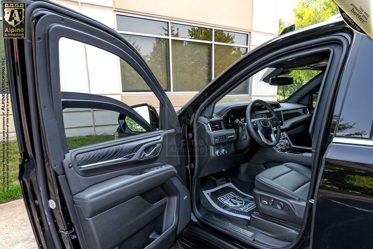 The interior of an armored GMC Yukon Denali XL with the driver's seat and passenger seat visible. Both doors are open, revealing a black and grey interior with leather.