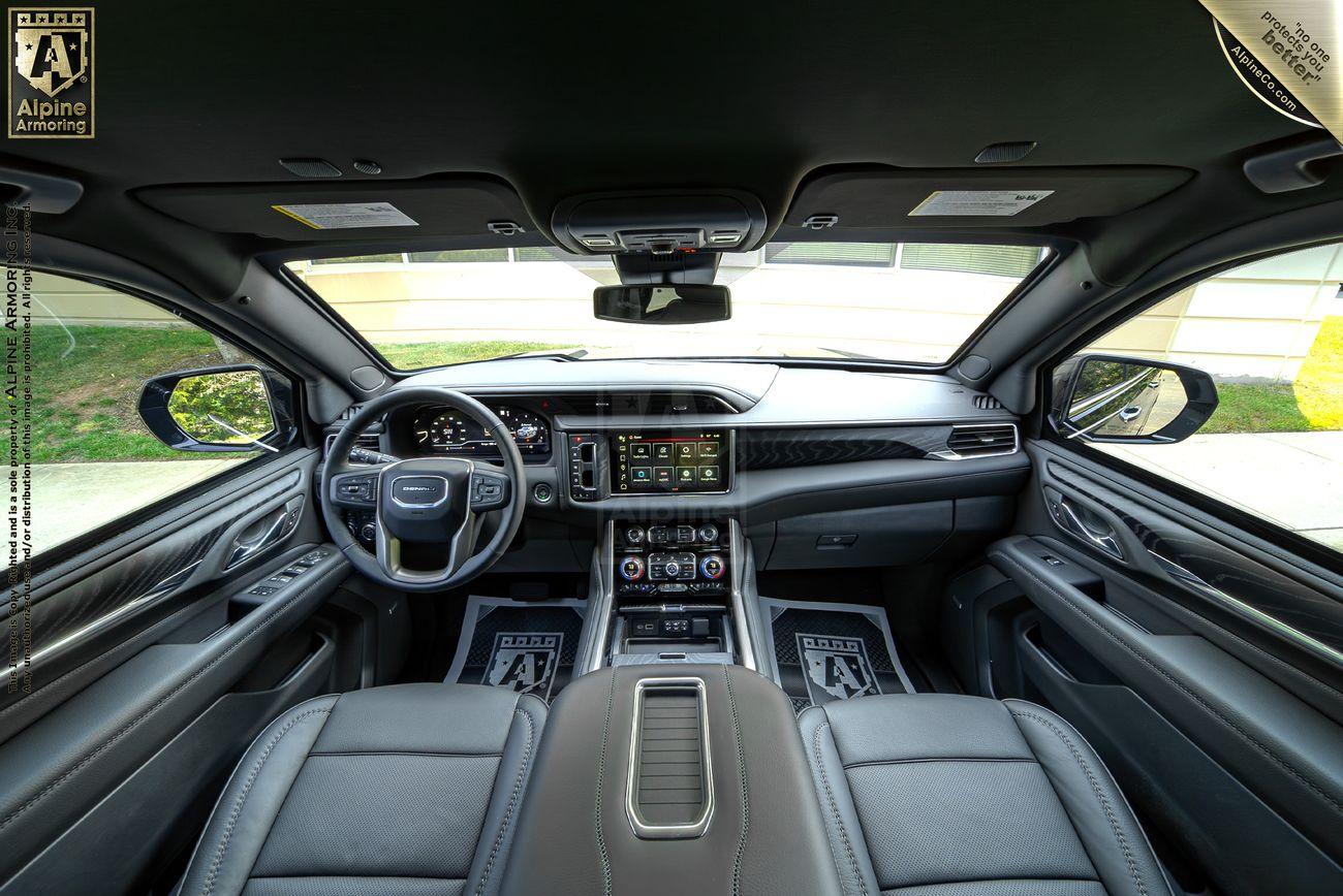 Interior of an armored GMC Yukon Denali XL SUV featuring a leather-wrapped steering wheel, large touchscreen display, and various control buttons on the dashboard and center console, with an Alpine Armoring logo visible.