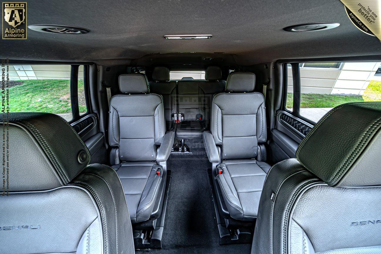 Interior view of a black armored GMC Yukon Denali XL with black leather seats arranged in three rows. The second row features captain’s chairs, and there are windows on both sides. A "Denali" logo is visible on one headrest.