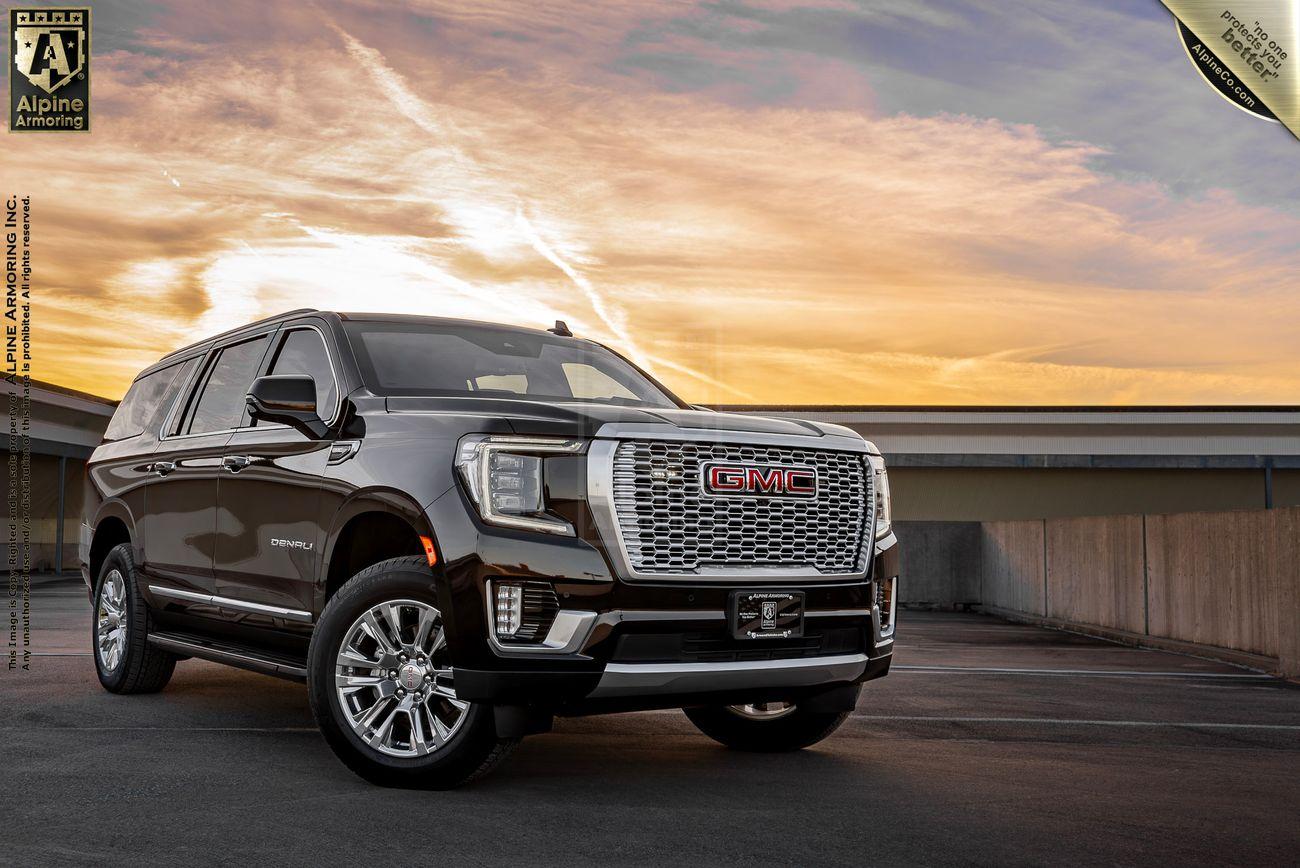 A black armored GMC Yukon Denali XL truck is parked on a rooftop parking lot at sunset with a building in the background. The sky is filled with orange and yellow clouds.