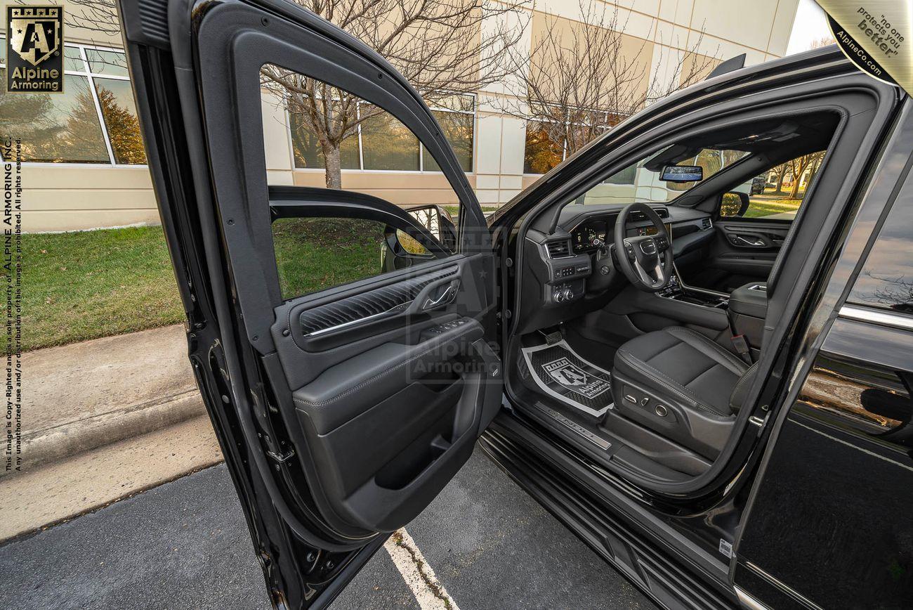 A black armored GMC Yukon Denali XL with the driver's side door open, revealing a clean and modern interior. An "Alpine Armoring" logo is visible on the door panel and floormat. The vehicle is parked near a building.