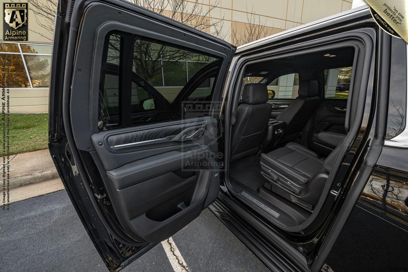 A rear view of a black bulletproof GMC Yukon Denali XL in a parking structure, showcasing its tail lights, license plate, and dual exhaust system. Branding for Alpine Armoring is visible on and above the vehicle.