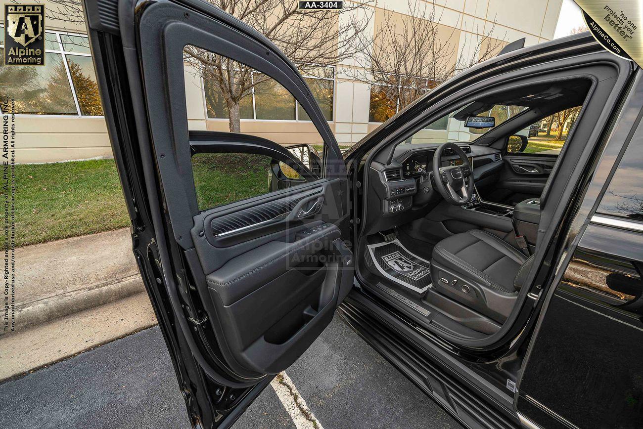 Open driver's side door of a black luxury GMC Yukon Denali XL, showing the interior with leather seats and a view of the dashboard. The car is parked outside a modern building.