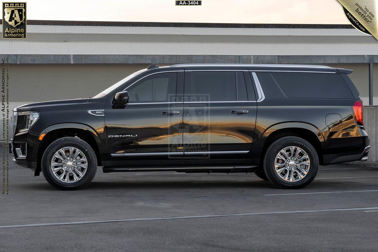 Driver side view of a black armored GMC Yukon Denali SUV parked in front of a building with beige and gray tones. The vehicle has chrome accents and polished wheels.