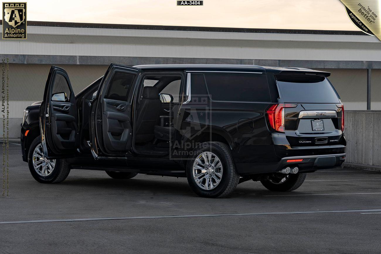 Black GMC Yukon Denali XL SUV with driver and rear passenger side doors open, parked in an empty lot with a building in the background. The Alpine Armoring logo is visible on the rear of the vehicle.