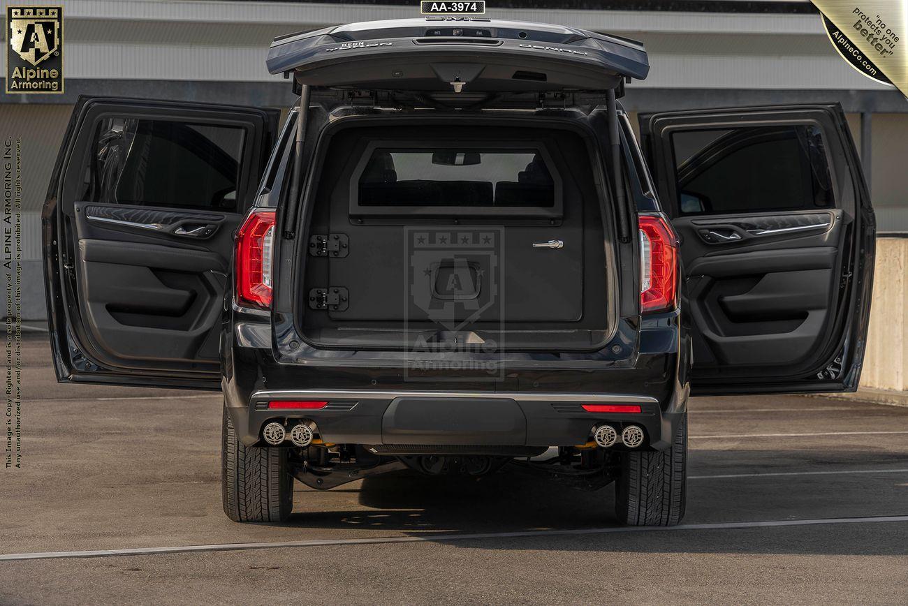 Rear view  of a black arnmored GMC Yukon Denali SUV, with the doors open, showing dual exhaust pipes, displayed in a parking area. 