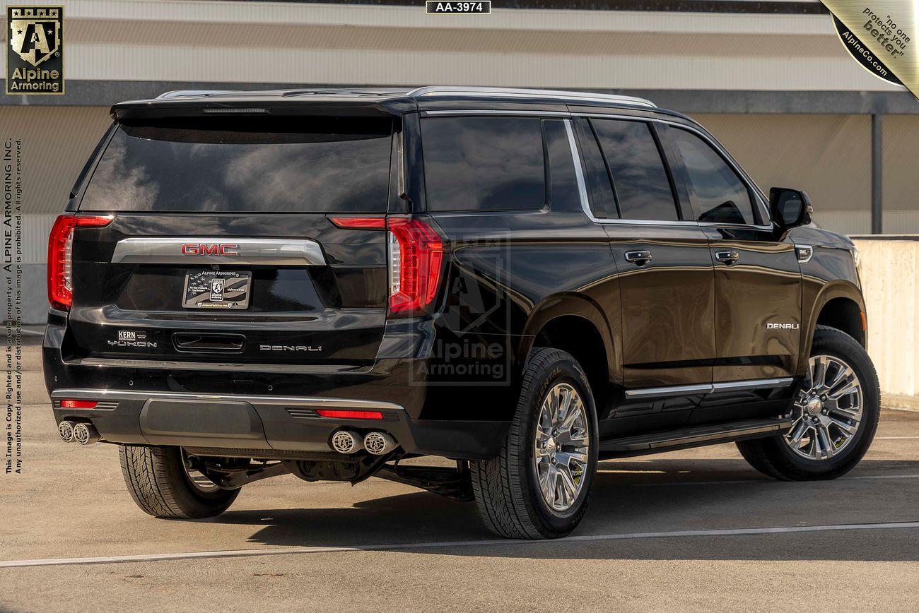 Rear view from the right side of a black arnmored GMC Yukon Denali SUV with dual exhaust pipes, displayed in a parking area. The vehicle has "Alpine Armoring" branding on the back.