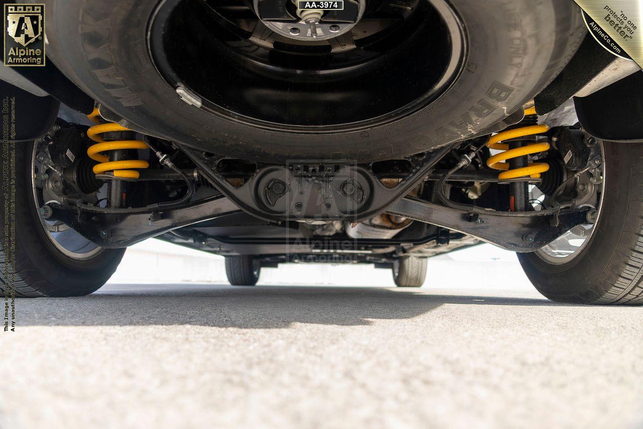 View of the underside of a black armored GMC Yukon Denali XL showing its suspension system with yellow springs and a spare tire mounted centrally. The ground is concrete.