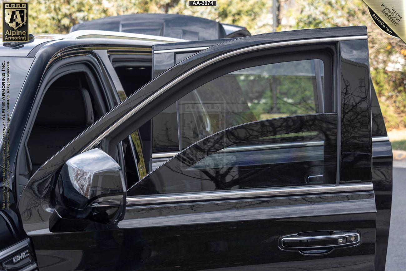 A close-up view of an open rear passenger door and closed front passenger door on a black GMC Yukon Denali XL with bulletproof glases, reflective windows and a logo decal visible on the upper left corner. Trees are seen in the background.