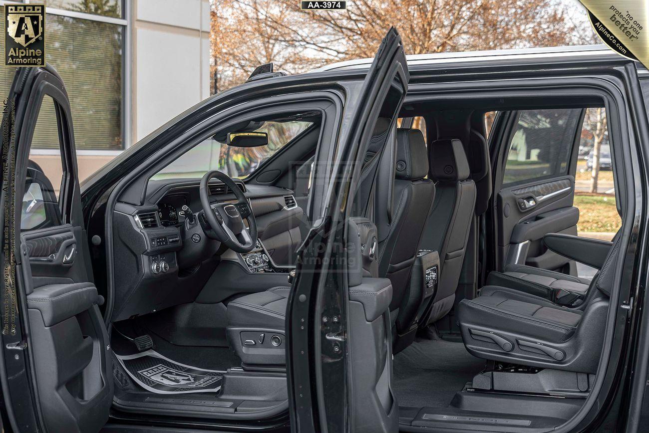 Image of a black armored GMC Yukon Denali XL SUV with its doors open showing a view of the interior, including the front and rear seats, dashboard, and steering wheel. The vehicle is parked outside near a building with large windows.