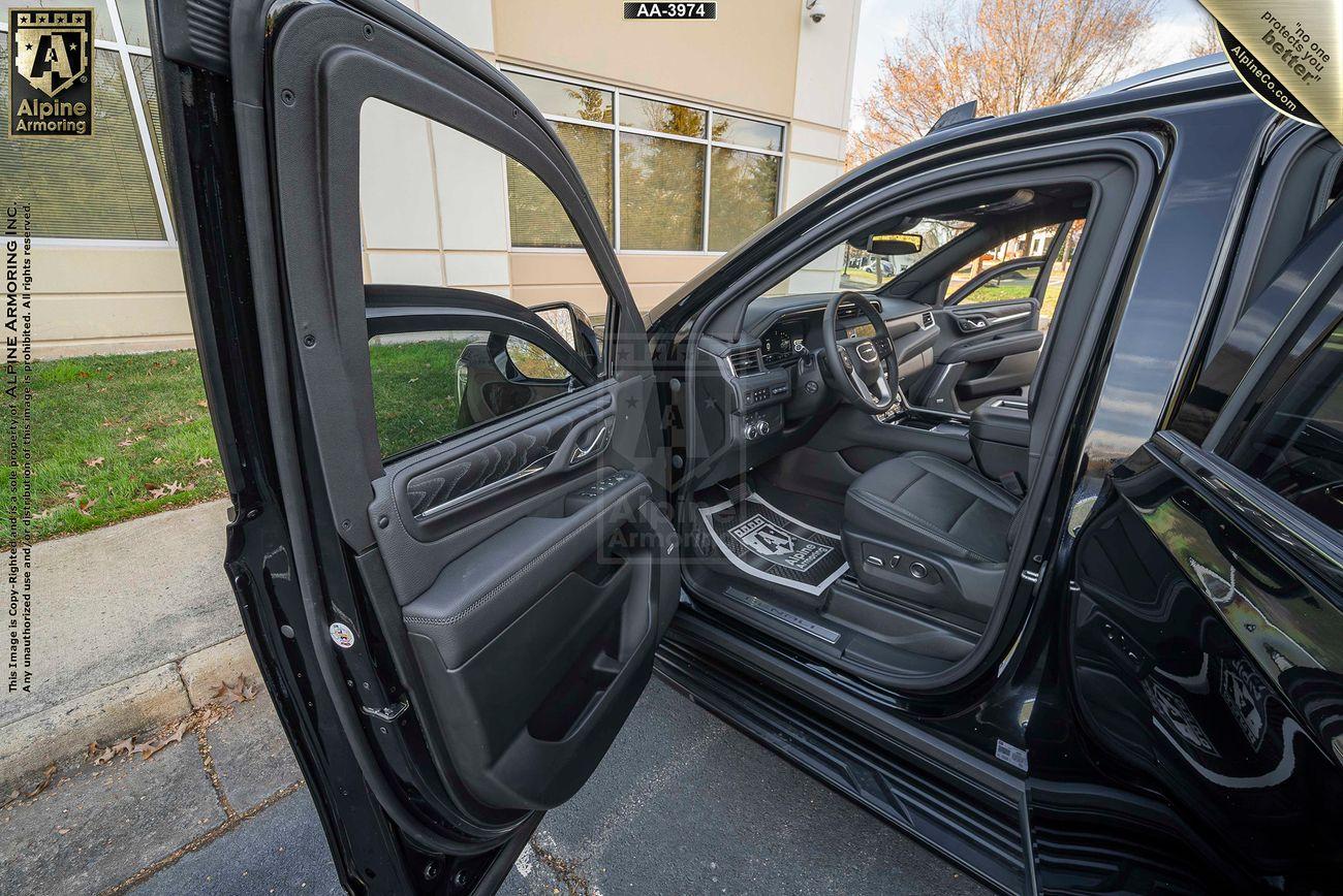 An armored  GMC Yukon Denali XL with its front driver-side door open, showing a clean and modern interior, parked outside a building.