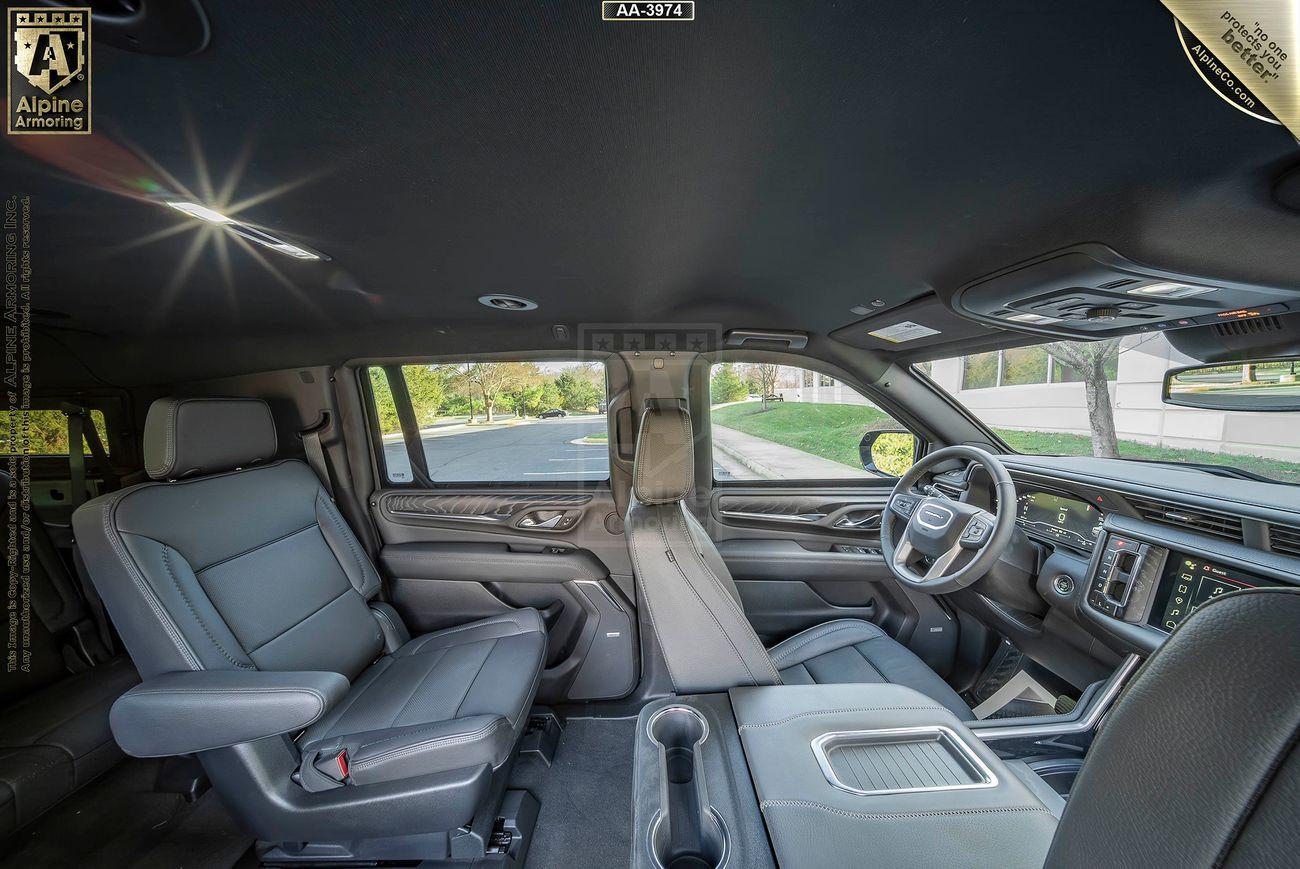 Interior of a bulletproof GMC Yukon Denali XL SUV showcasing front and rear seats, a steering wheel, dashboard, and a mounted center display. The seats are upholstered in grey material, and natural light is entering through the windows.