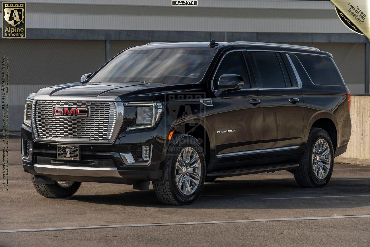 Front view from the left side of a black arnmored GMC Yukon Denali SUV with dual exhaust pipes, displayed in a parking area. The vehicle has "Alpine Armoring" branding on the back.