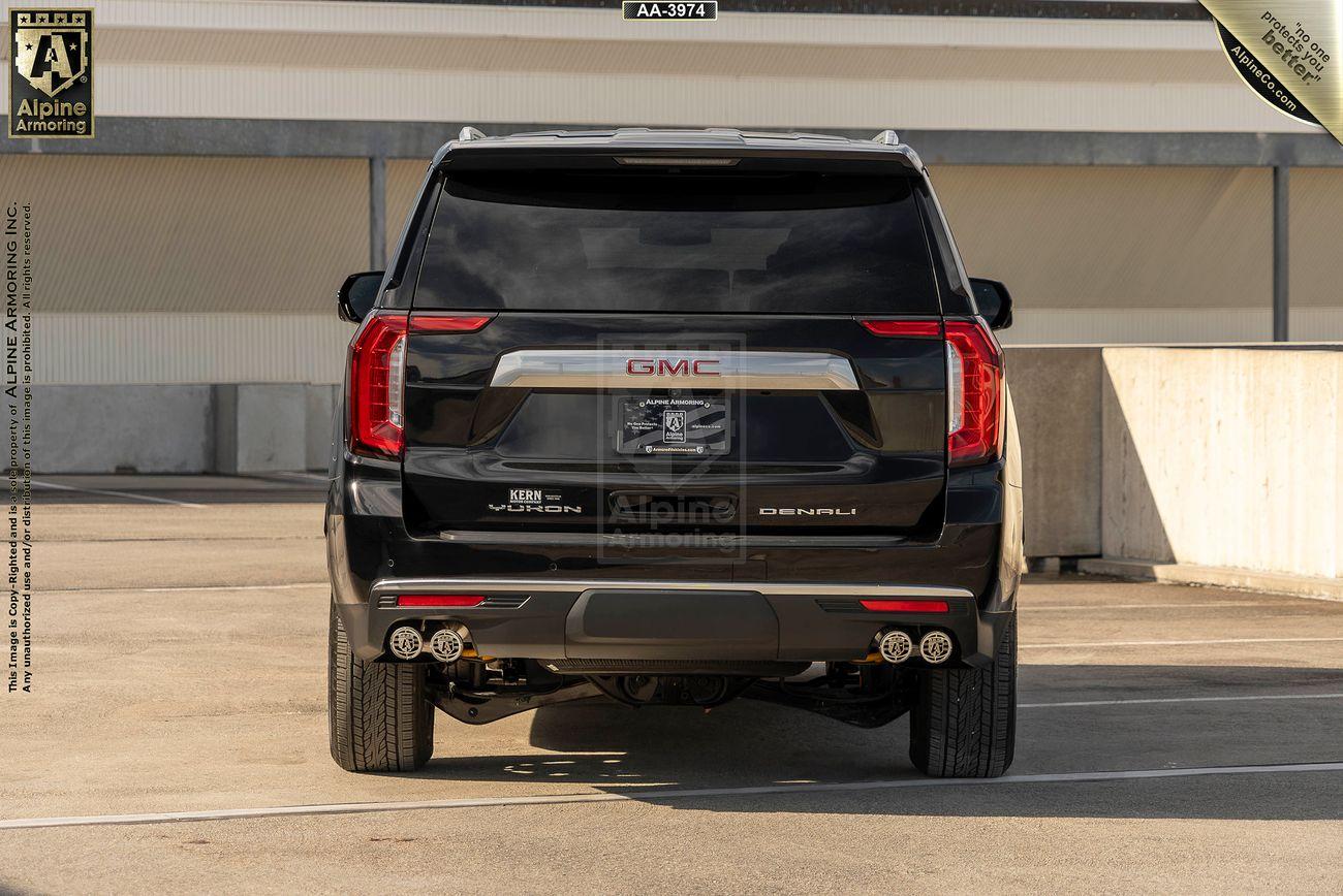 Rear view of a black arnmored GMC Yukon Denali SUV with dual exhaust pipes, displayed in a parking area. The vehicle has "Alpine Armoring" branding on the back.