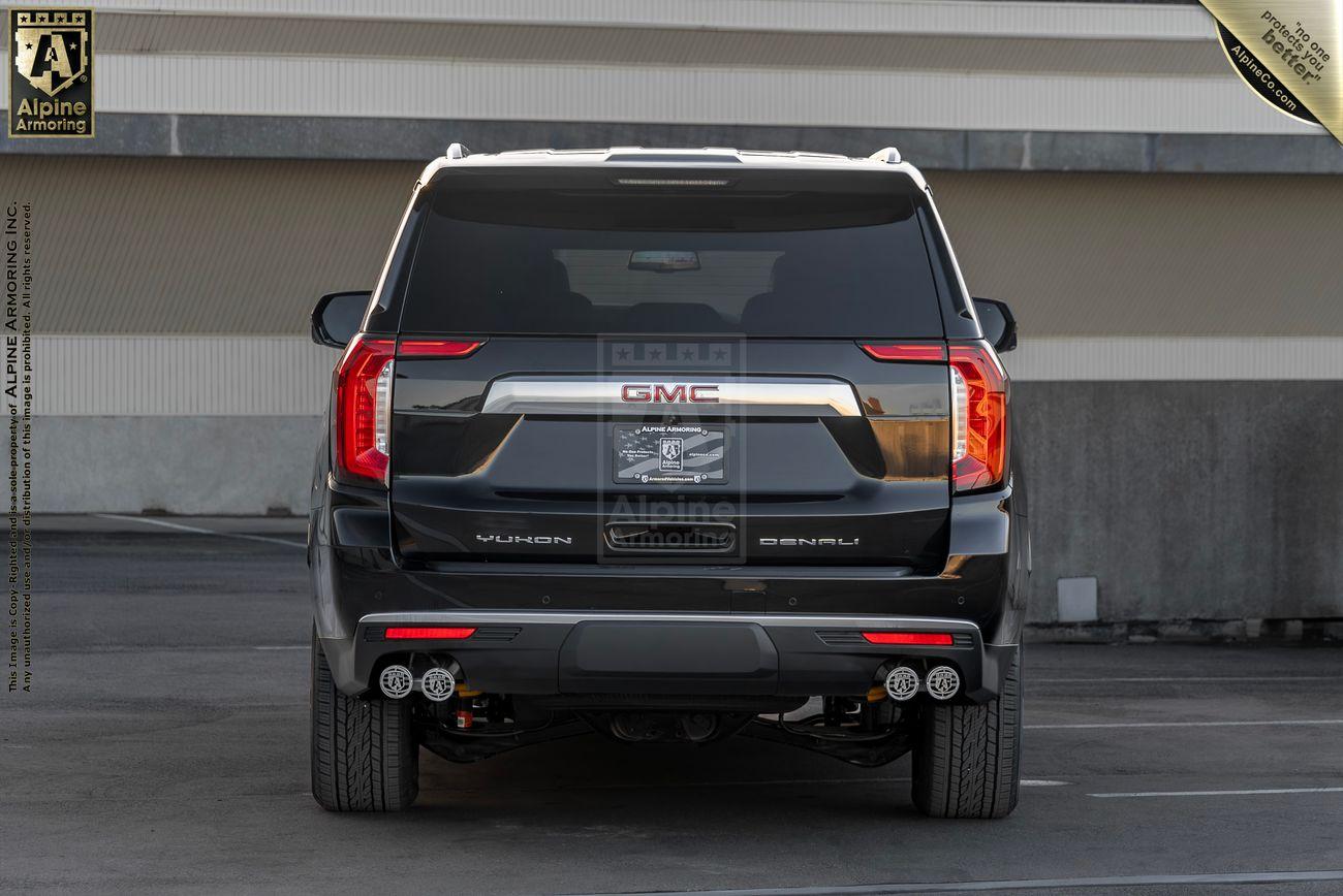 A rear view of a black GMC Yukon Denali XL SUV in a parking structure, showcasing its tail lights, license plate, and dual exhaust system. Branding for Alpine Armoring is visible on and above the vehicle.