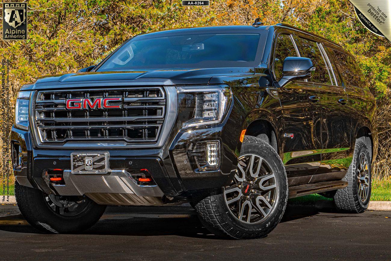 A black armored GMC Yukon XL AT4 parked on a road with a backdrop of greenery and trees. The vehicle features chrome detailing and off-road tires.