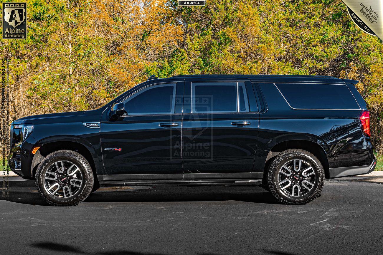 A black armored GMC Yukon XL AT4 SUV is parked on pavement in front of green foliage. The vehicle has a logo on the rear side door and is equipped with black rims and tinted windows.