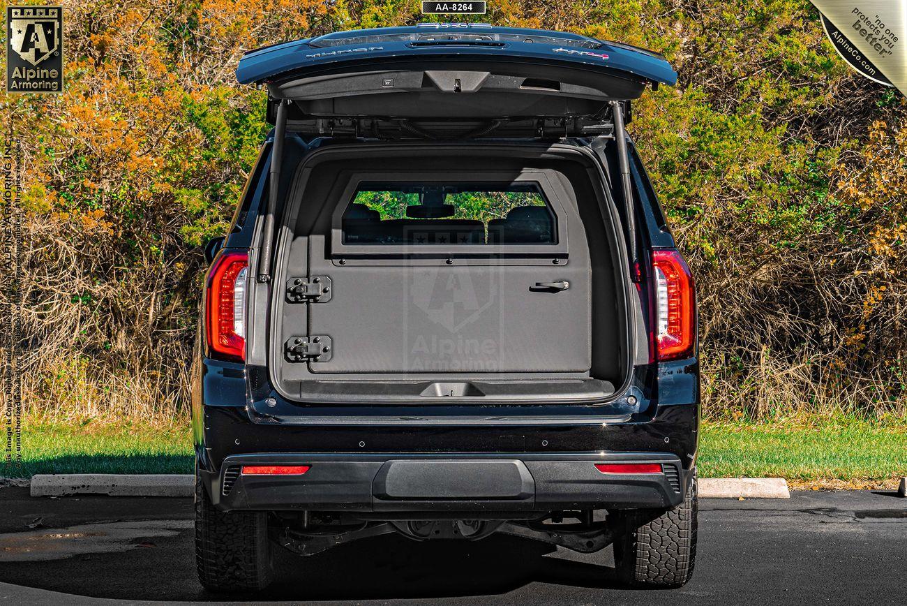 Rear view of an GMC Yukon XL AT4 SUV with its tailgate and rear doors open, revealing the empty cargo area. The vehicle is parked on a paved area with greenery in the background.