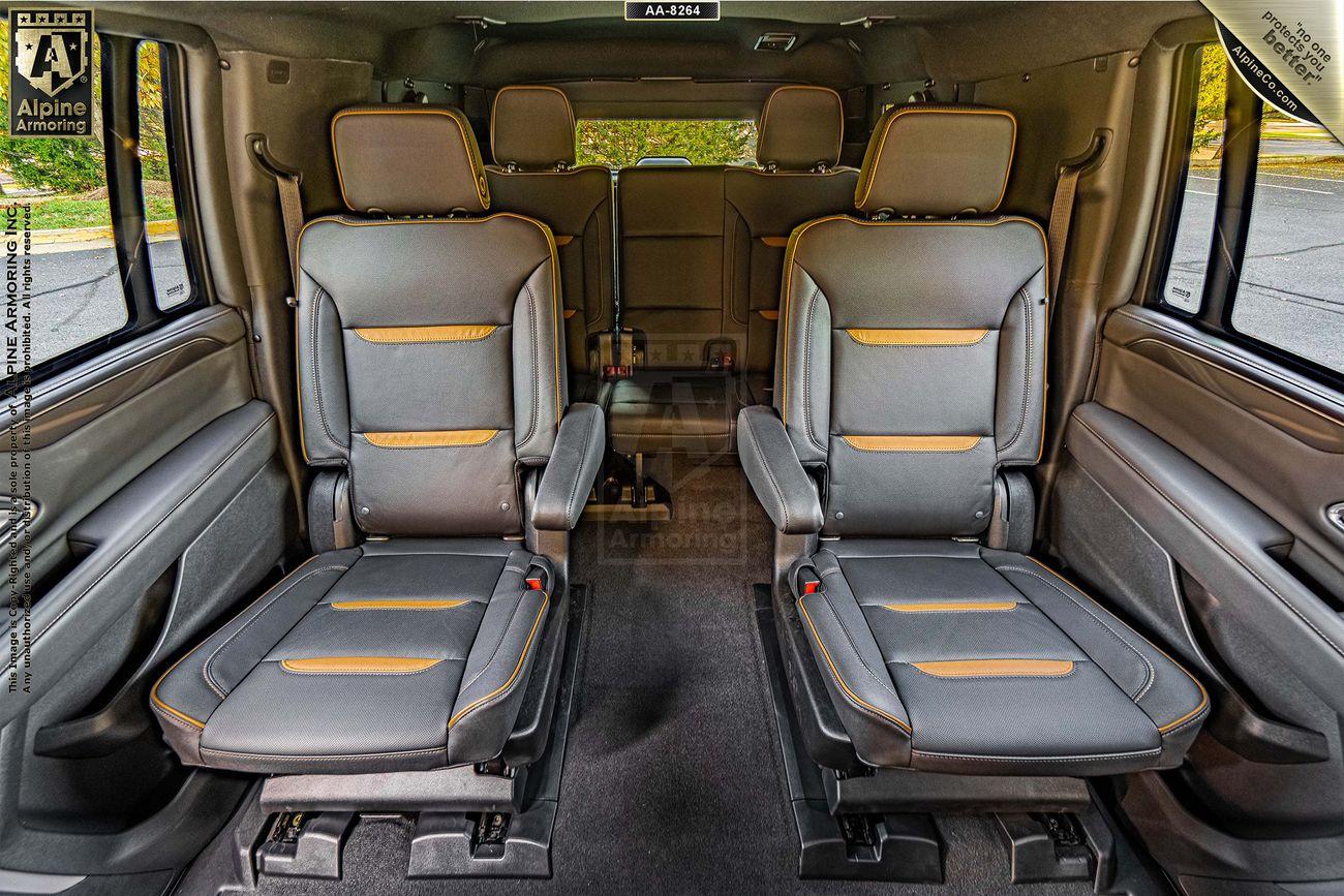 Interior view of an armored GMC Yukon XL featuring four black leather seats arranged in two rows, with gold accents. The seats are designed with armrests and headrests, set against a backdrop of windows and a carpeted floor.