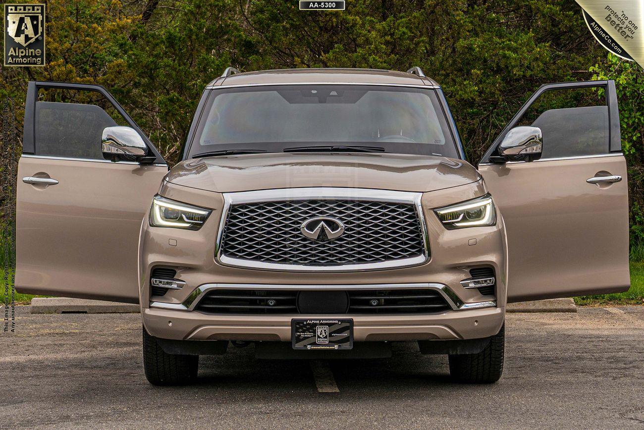 Front view of a beige INFINITI QX80 SUV with both front doors open, parked on a road with greenery in the background.