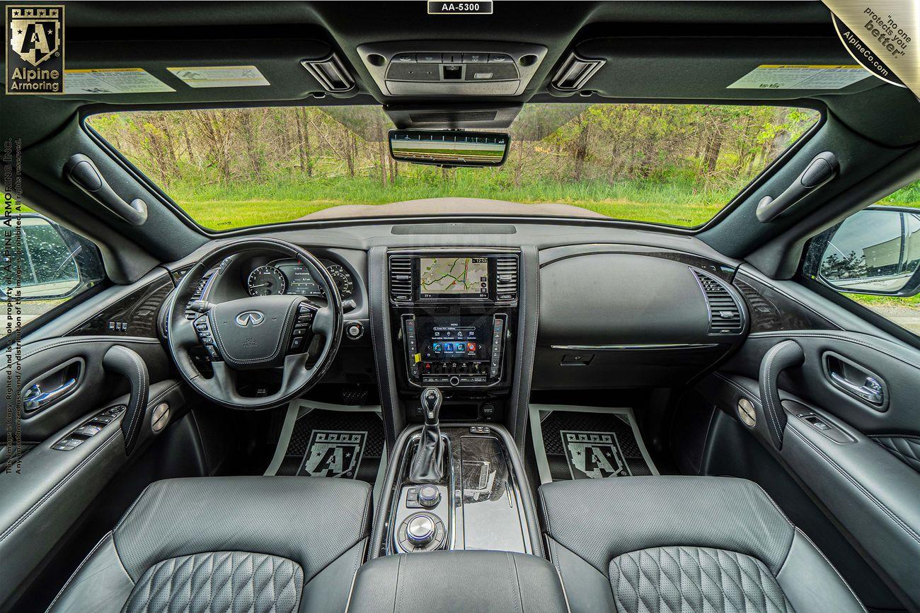 Interior view of a INFINITI QX80 car showcasing the dashboard, steering wheel, center console with touchscreen display, and luxurious seats with detailed stitching. Large sunroof and green landscape outside.