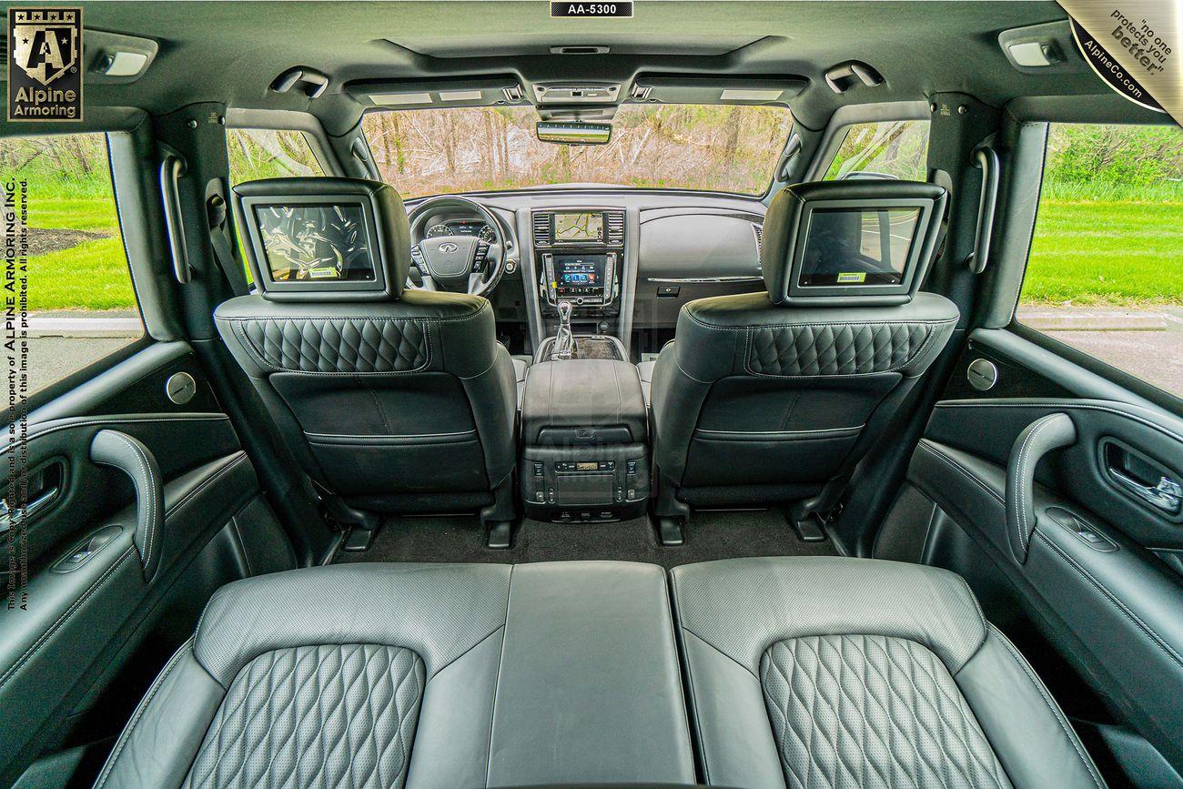 Interior view of an armored INFINITI QX80 featuring black leather seats, a central console, digital screens on the back of the front seats, and a clear view through the rear window showing a green landscape.