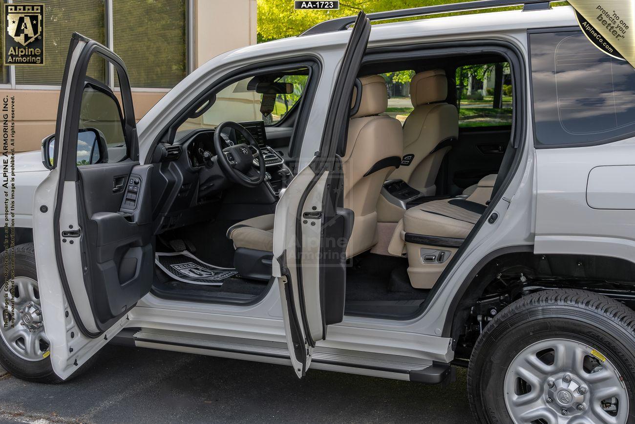 A white Armored Toyota Land Cruiser 300 SUV with all four doors open, revealing a beige leather interior with front and rear seats and a visible dashboard. The vehicle is parked outside a building.