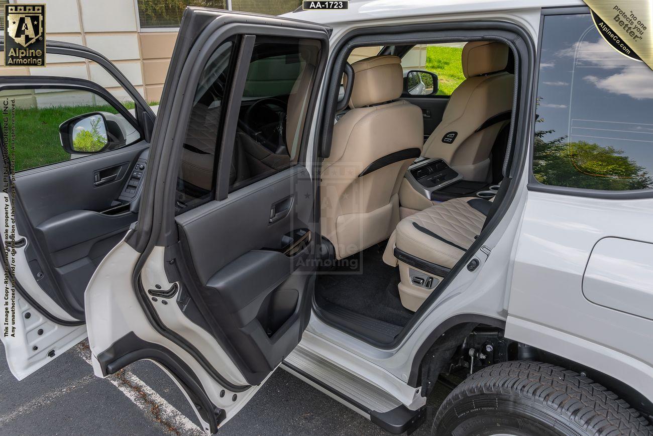 The image shows the backseat and front seat of a white Armored Toyota Land Cruiser 300  with both side doors open. The interior is cream-colored, with a center console visible in the backseat.