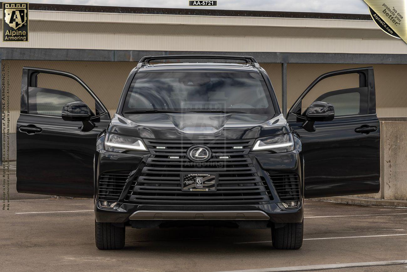 A black armored Lexus LX600 Luxury SUV with its front doors open, displayed in front of a concrete building. The vehicle has a license plate and is showcased under a "Alpine Armoring" sign.